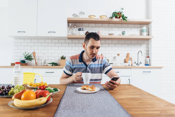 Nuori mies käyttää älypuhelinta syödessään aamiaista kotona
. - Valokuva, kuva