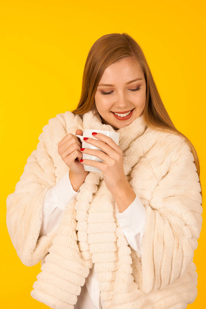 Beautiful woman in a fur winter jacket drinks tea over yellow ba - Fotoğraf, Görsel