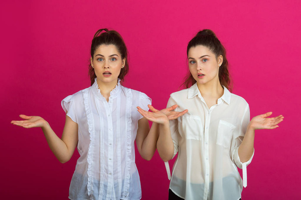two beautiful young sisters twins in a good mood in white blouses on a pink background with a facial expression - 写真・画像