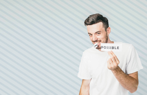 Attractive and posistive guy is standing at the white wall and holding a piece of paper in his hand and rest of it with his teeth. It was said Impossible but right now it says Possible on that paper. - Foto, afbeelding