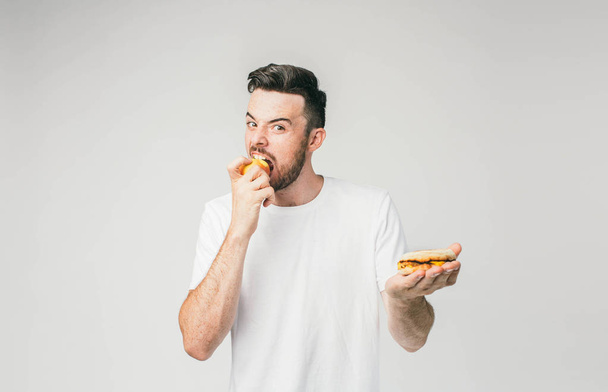 A picture of dark-haired guy looking straight in front of himself and eating an apple with the right hand. Also he is holding a burger in his left hand. - Photo, image