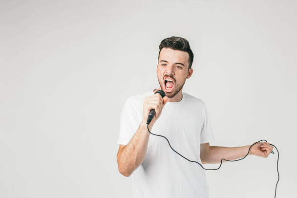 Another picture of a guy in white t-shirt holding a mic in right hand and a cord in his left hand and singing aloud and very confident. He is sure his singing is amazing. - Photo, Image