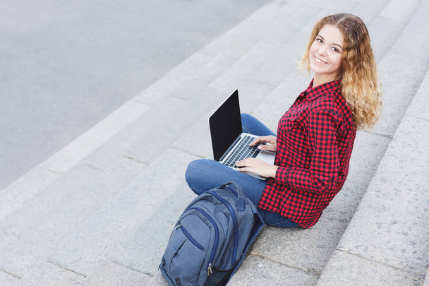 Smiling student girl sitting on the stairs using laptop outdoors - Valokuva, kuva