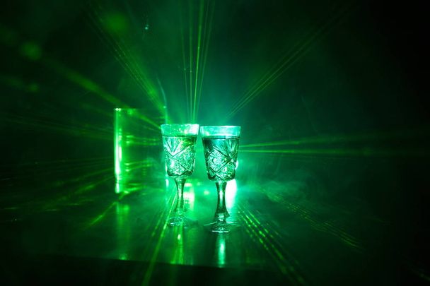 Dos vasos de vodka con botella sobre fondo oscuro niebla estilo club con luces brillantes (Láser, Stobe) Multi color. Club bebidas decoración temática. Espacio vacío
 - Foto, Imagen