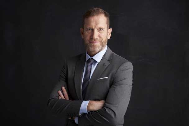 Portrait of middle aged wrinkled businessman with arms crossed wearing suit while posing at dark background. - Fotografie, Obrázek