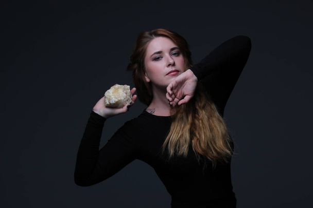 portrait of young beautiful red hair woman in black dress holding sea stone on dark background - Photo, Image