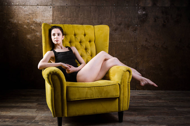 A young beautiful, sexy Caucasian woman with thin figure and long bare legs, barefoot posing reclining on yellow armchair in the interior against dark wooden wall. Dressed in a black classic swimsuit - Foto, Imagem