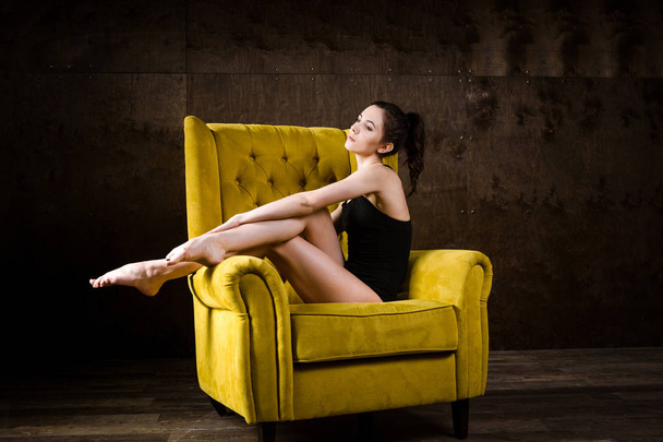A young beautiful, sexy Caucasian woman with thin figure and long bare legs, barefoot posing reclining on yellow armchair in the interior against dark wooden wall. Dressed in a black classic swimsuit - Foto, Imagem