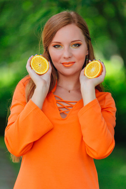 Portrait of pretty red hair woman holding two slices of juicy delicious oranges at summer green park. - Foto, Imagen