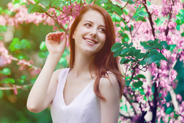 Młoda ruda dziewczyna w białej sukni stojącej w pobliżu drzew i krzewów w parku. Sezon wiosenny  - Zdjęcie, obraz