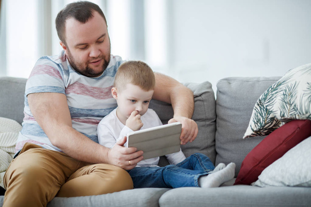 Picture of father with son with tablet sitting on gray sofa - Foto, Imagen