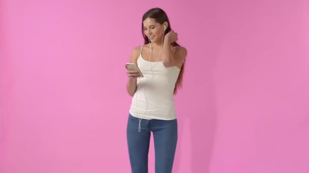 A girl in jeans and a T-shirt listening to music in headphones on a pink background - Video