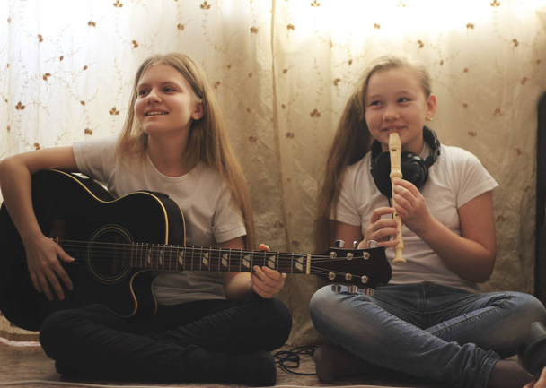 Two female teens playing musical instruments, guitar and flute, sitting on the floor at home - Photo, Image