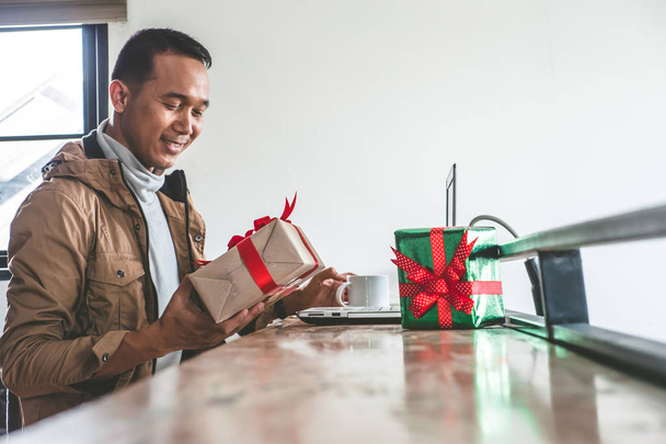 Happy man holding gift box and using laptop on the table. Asian man preparing presents for christmas holiday. - Photo, Image