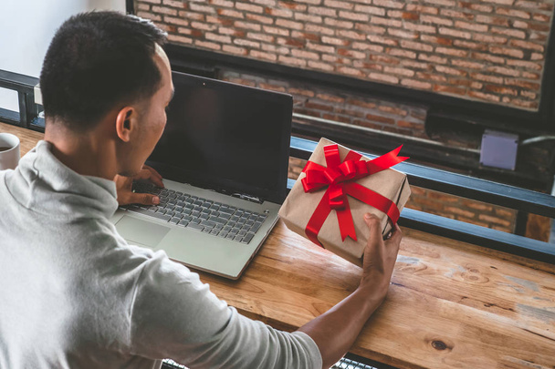 Happy man holding gift box and using laptop on the table. Asian man preparing present for christmas holiday. - Photo, Image