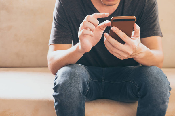 Close up of casual young man sitting on sofa and usig a smart phone at home. - Photo, Image