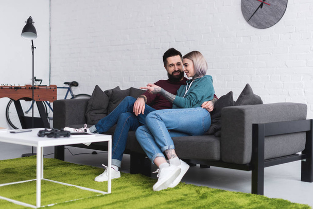 smiling tattooed couple hugging on sofa in living room - Фото, изображение