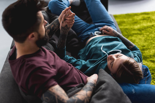 tattooed couple lying on sofa and holding hands - Zdjęcie, obraz