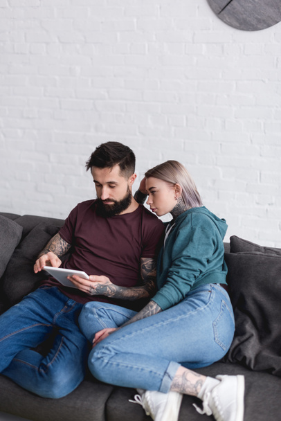 tattooed couple looking at tablet on sofa in living room - Фото, зображення