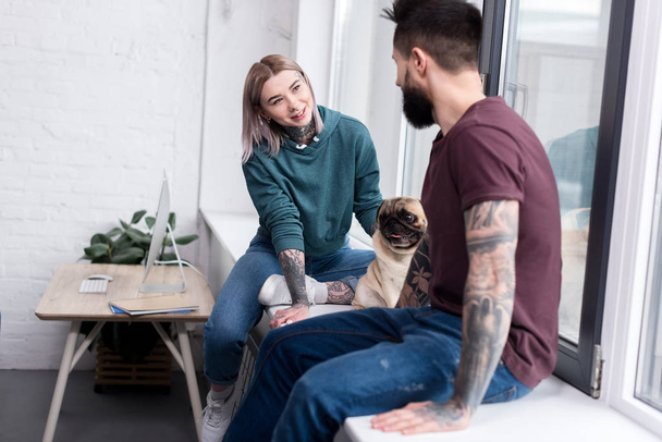 happy tattooed couple sitting on windowsill with pug at home - Foto, Imagen
