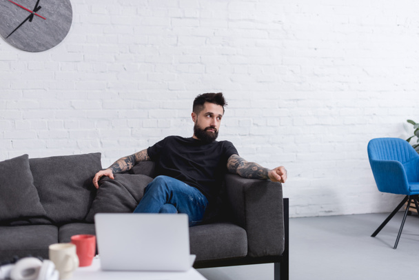 tetovált szép ember ül a kanapén, és keres el otthon - Fotó, kép