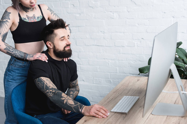 cropped image of tattooed girlfriend touching boyfriend near table with computer at home - Foto, Imagem