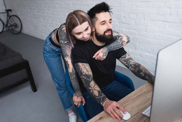 tattooed girlfriend hugging boyfriend while he using computer at home - Foto, afbeelding