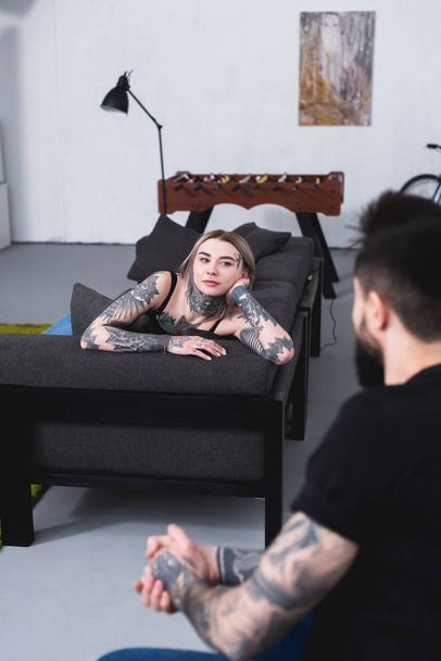 tattooed girlfriend lying on sofa and looking at boyfriend at home - Фото, зображення