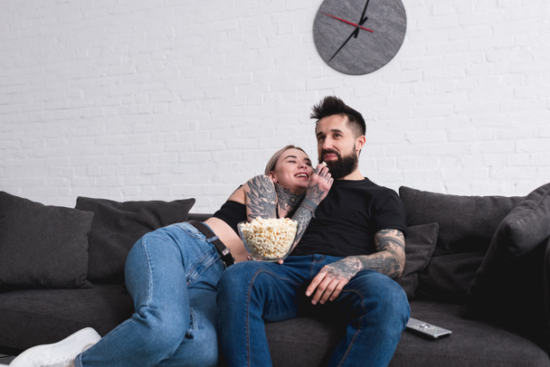 tattooed couple eating popcorn on sofa at home - Фото, зображення