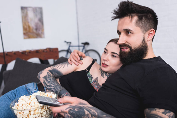 affectionate tattooed couple watching tv with popcorn at home - Foto, afbeelding
