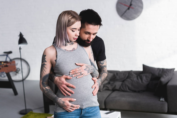 happy tattooed boyfriend hugging pregnant girlfriend from back in living room - Photo, image