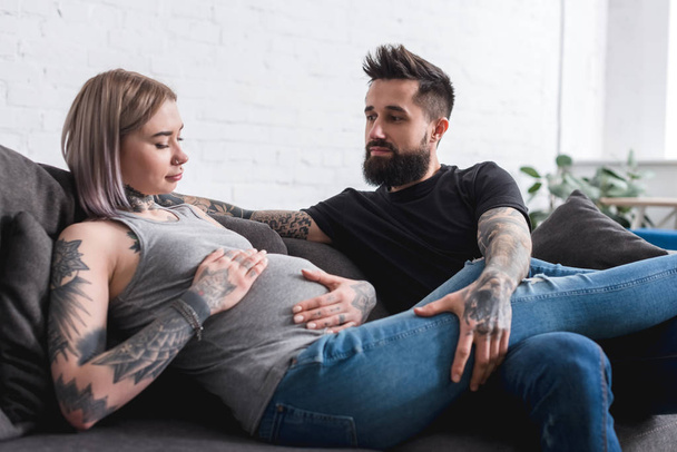 tattooed pregnant girlfriend touching abdomen near boyfriend at home - Фото, зображення