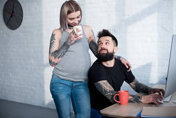 tattooed pregnant girlfriend drinking tea and hugging boyfriend at home - Fotó, kép