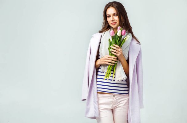 Fashion photo of a beautiful young woman with tulips in her hand .she dressed in a beautiful coat, scarf, pants and T-shirt with stripes.Spring concept. March 8. beautiful girl in stylish clothes - Photo, Image