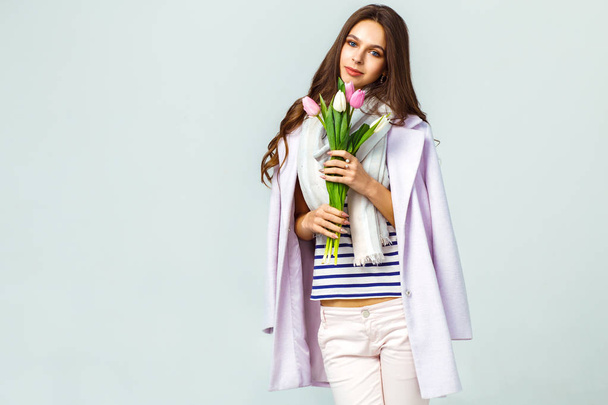 Fashion photo of a beautiful young woman with tulips in her hand .she dressed in a beautiful coat, scarf, pants and T-shirt with stripes.Spring concept. March 8. beautiful girl in stylish clothes - Photo, image