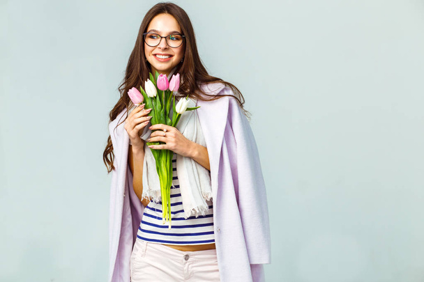 Fashion photo of a beautiful young woman with tulips in her hand .she dressed in a beautiful coat, scarf, pants and T-shirt with stripes.Spring concept. March 8. beautiful girl in stylish clothes - Zdjęcie, obraz