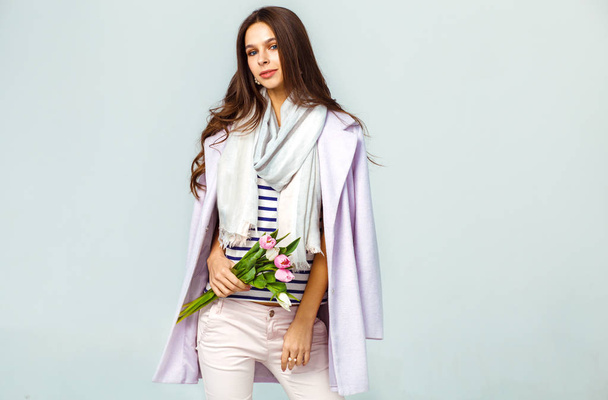 Fashion photo of a beautiful young woman with tulips in her hand .she dressed in a beautiful coat, scarf, pants and T-shirt with stripes.Spring concept. March 8. beautiful girl in stylish clothes - Fotó, kép