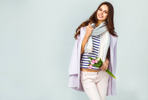 Fashion photo of a beautiful young woman with tulips in her hand .she dressed in a beautiful coat, scarf, pants and T-shirt with stripes.Spring concept. March 8. beautiful girl in stylish clothes - Foto, Bild
