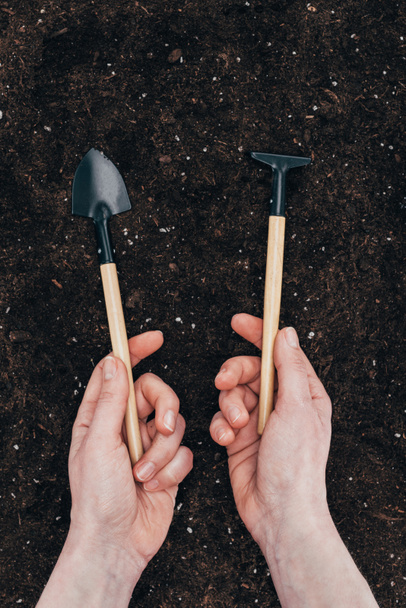 recortado tiro de manos humanas sosteniendo pequeñas herramientas de jardinería sobre el suelo
 - Foto, imagen
