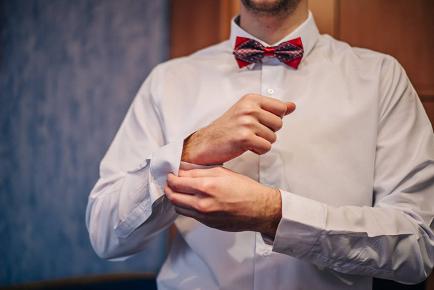 o noivo prende um botão na manga de uma camisa com uma borboleta vermelha em seu pescoço em vez de um empate no dia do casamento
 - Foto, Imagem