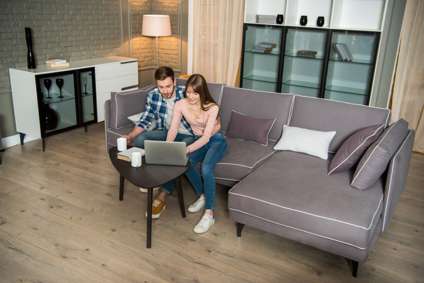 Vysoký úhel pohled šťastný pár sedí na gauči a používat přenosný počítač v obývacím pokoji v moderním designu - Fotografie, Obrázek