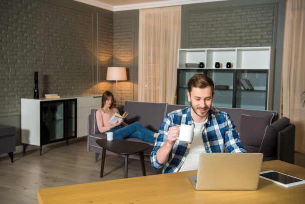 Muž s šálkem v ruce pomocí přenosného počítače, zatímco žena čte knihu na gauči v obývacím pokoji v moderním designu - Fotografie, Obrázek