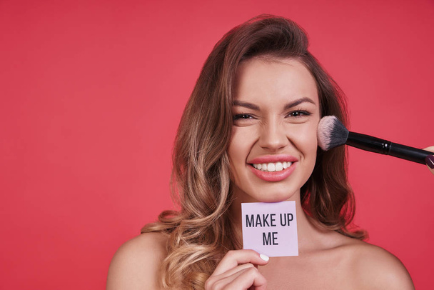 Beautiful young woman smiling and applying make up with brush, holding paper poster  - Foto, afbeelding