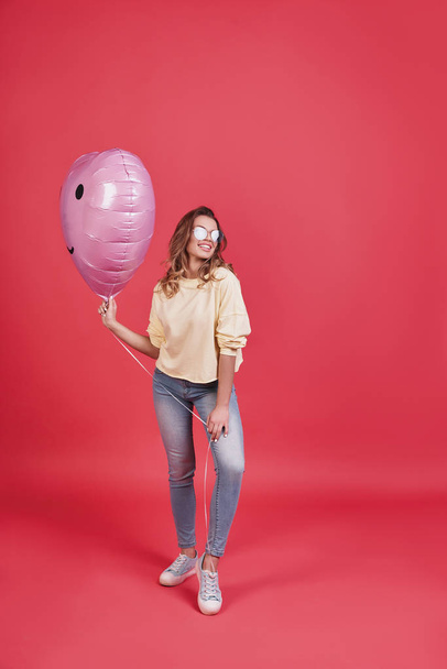 Full length of attractive young woman holding balloon and standing against pink background in studio - Photo, Image