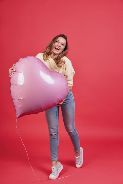 playful woman holding heart shaped balloon and smiling against pink background - Фото, изображение