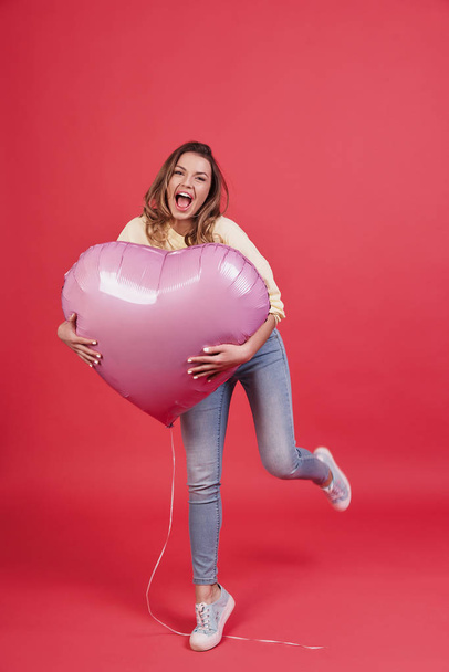 playful woman holding heart shaped balloon and smiling against pink background - Foto, imagen