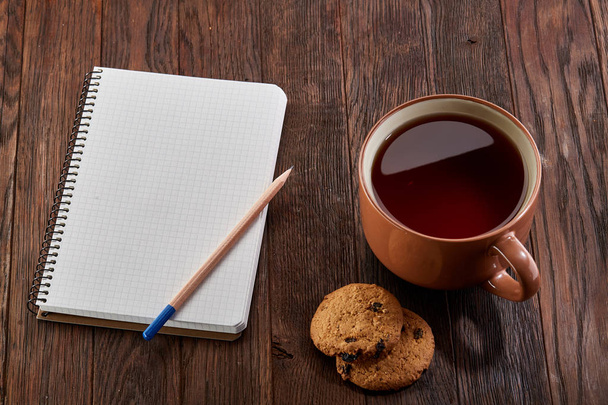 Cup of tea with cookies, workbook and a pencil on a wooden background, top view - Photo, image