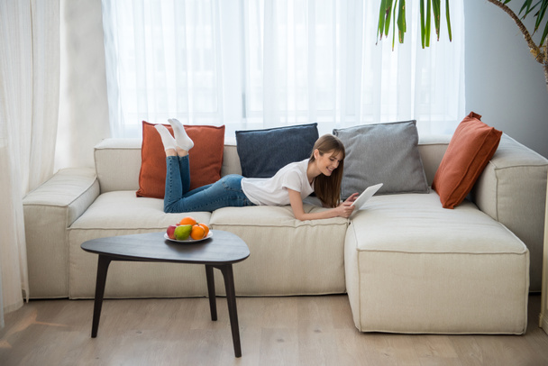 Widok z boku z młoda kobieta, leżąc na kanapie i za pomocą cyfrowego tabletu w salonie z nowoczesnym wnętrzem - Zdjęcie, obraz