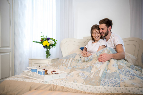 Happy couple sitting on bed with breakfast on tray in cozy bedroom - Photo, Image