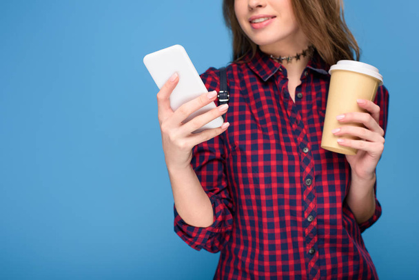 obcięty widok młodej dziewczyny z kawą do go za pomocą smartfona, na niebieskim tle - Zdjęcie, obraz